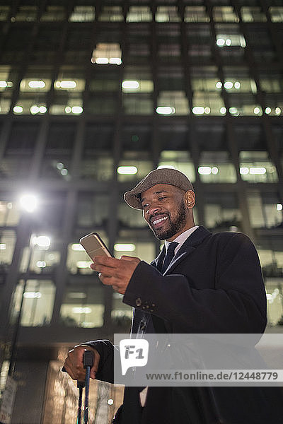 Smiling businessman with smart phone standing below highrise building at night