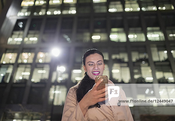 Smiling businesswoman using smart phone below urban highrise at night