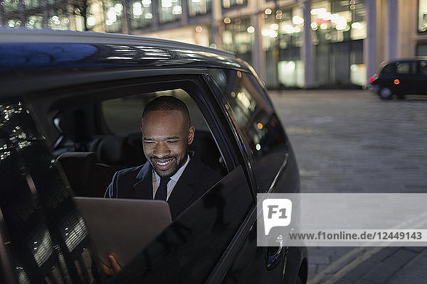 Smiling businessman using laptop in crowdsourced taxi at night