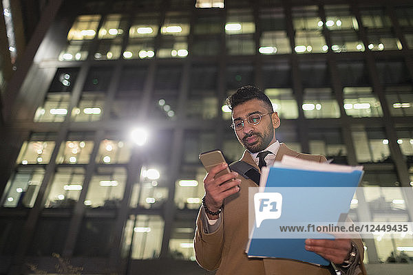 Businessman with paperwork and smart phone below urban highrise at night