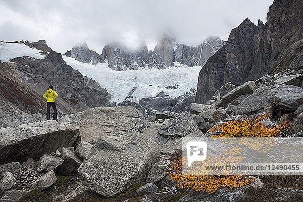 Female hiker with a hand-on-hip pose standing confident on a boulder and looking towards high peaks shrouded in clouds and a glacier below them