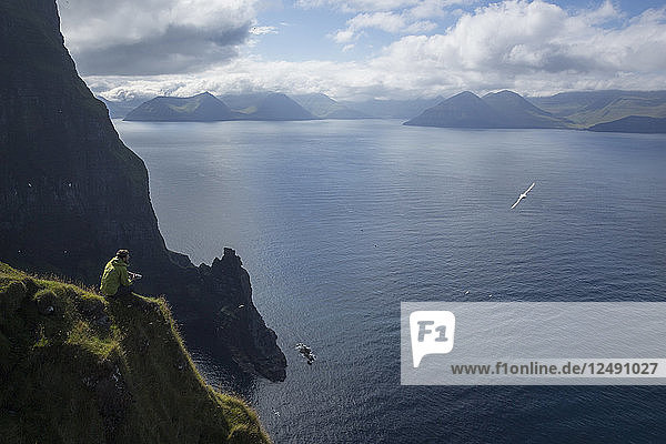 Männlicher Wanderer sitzt auf einem Felsvorsprung über dem Meer und blickt auf die fernen Berge  während eine Möwe vorbeifliegt.