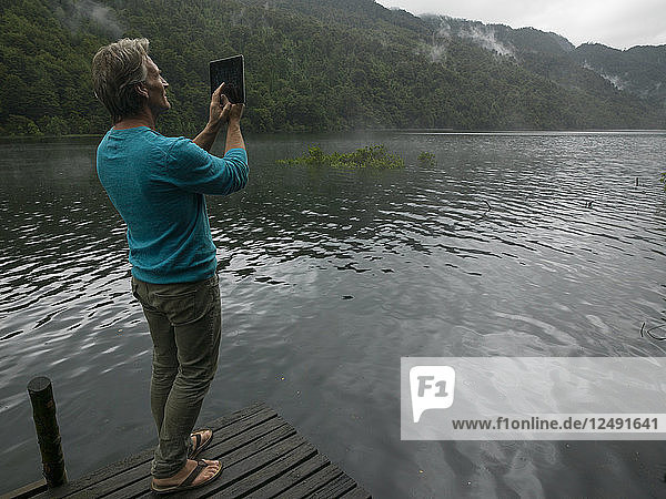 Mature man takes photo with tablet on dock in mountain lake
