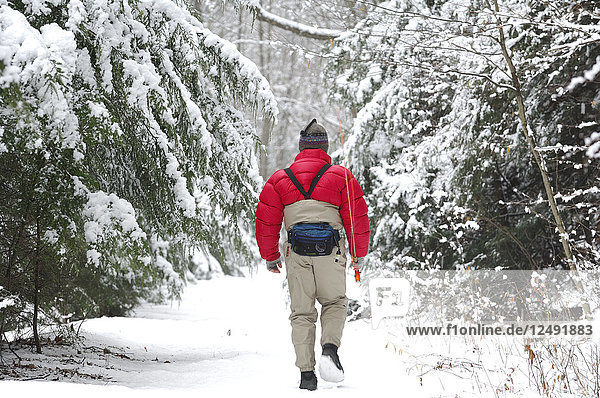 Ein Mann geht an einem kalten  verschneiten Wintertag mit seiner Angelausrüstung durch den Wald  um einen guten Platz zum Fliegenfischen zu finden.