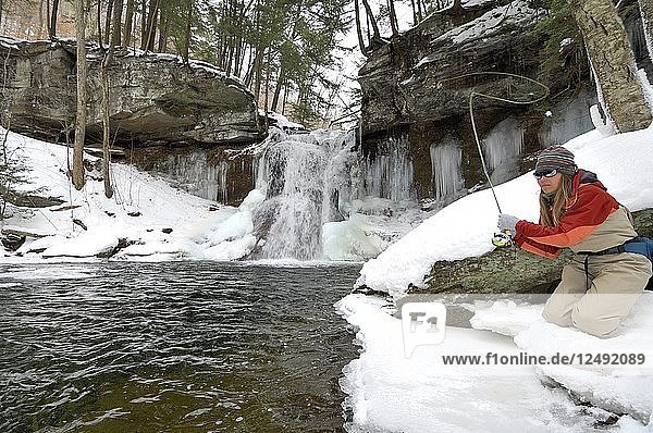 Eine junge Frau beim Fliegenfischen an einem verschneiten  kalten Wintertag. Sie angelt am Oberlauf eines Baches mit einem Wasserfall  an dem sich Eiszapfen gebildet haben.
