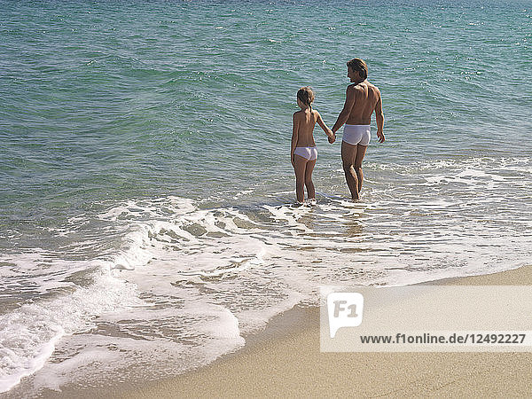 Vater und Tochter halten sich an den Händen und gehen vom Ufer ins Meer