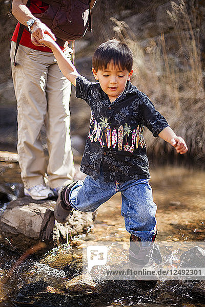 A four year old Japanese American boy crosses a stream while hiking along with his mother who carries her 4 month old baby  Hewlett Gulch Trail  Colorado.