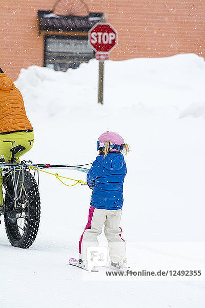 Eine Mutter zieht ihre Tochter mit Skiern auf einem Fahrrad mit großen Reifen durch die verschneiten Straßen von Silverton  Colorado.