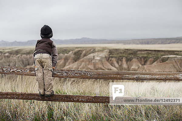 Ein 4 Jahre alter japanisch-amerikanischer Junge  der als Forscher mit Hut und Weste gekleidet ist  erkundet den Badlands-Nationalpark in South Dakota  während er auf dem Geländer eines Holzzauns steht und das Land erforscht.