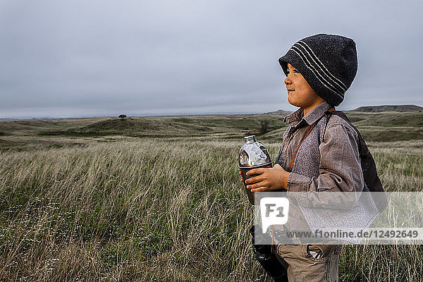 A 4 year old Japanese American boy dressed as an explorer with a hat and vest surveys the land (grasslands and prairies) and holds his old metal canteen in Badlands National Park  South Dakota.