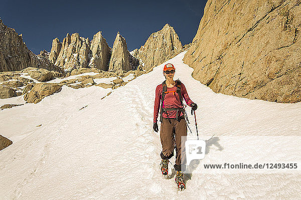 Eine 30-jährige Frau in Bergsteigerkleidung lächelt  während sie ein steiles Schneefeld hinabsteigt  hinter dem sich zackige Granitgipfel erheben.