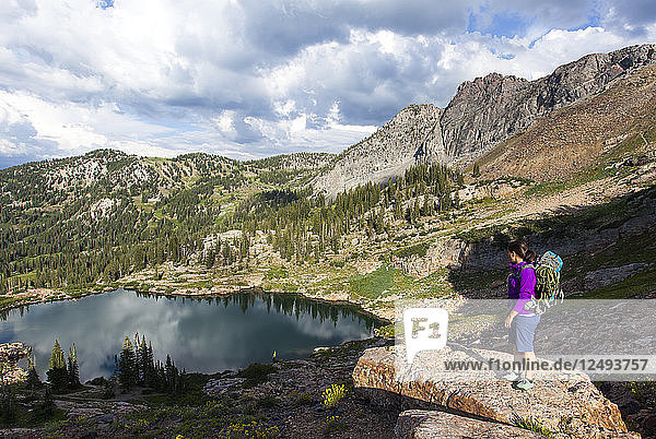 A women stands on a rocking outcrop and over looks Cecret Lake while hiking down from rock climbing on a summer evening.
