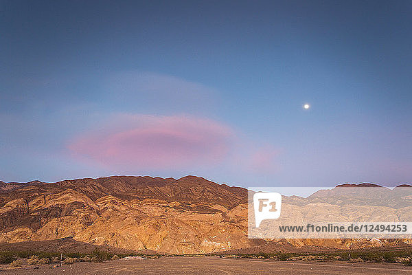 Der Mond geht an einem dunkelblauen Himmel mit einer rosafarbenen Wolke auf  die über einem Wüstengebirge hängt  das von der untergehenden Sonne orange beleuchtet wird.