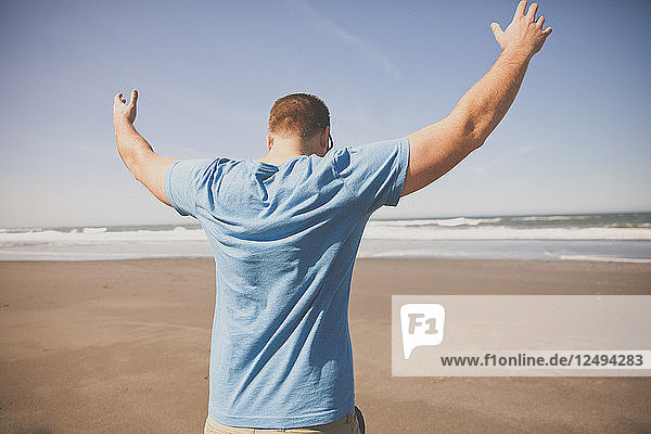 Ein Mann hebt beide Hände in die Luft  während er an einem Sandstrand an der Küste von Oregon spazieren geht.