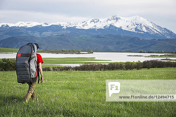 Young male hikes through grassy field towards a lake with an inflatable paddleboard in the mountains