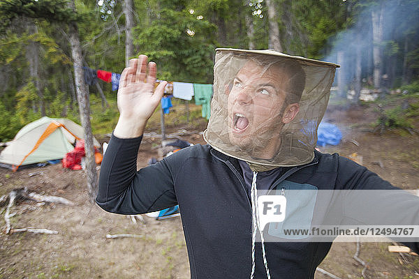 A man swats at mosquitoes while wearing a mosquito head net at his campsite.