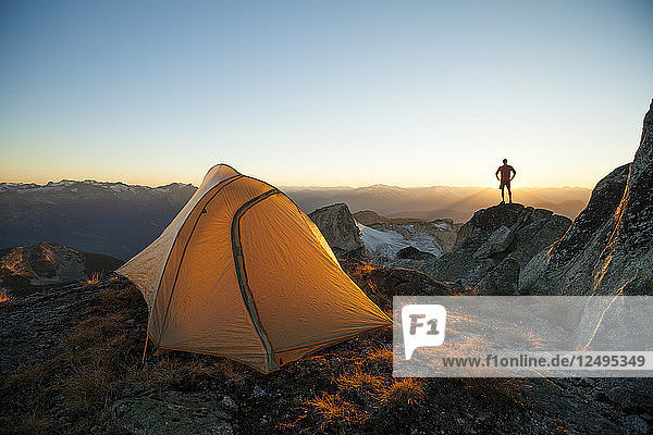 A silhouetted hiker stands on a rock bluff while camping on the summit of Saxifrage Peak  Pemberton  BC  Canada.
