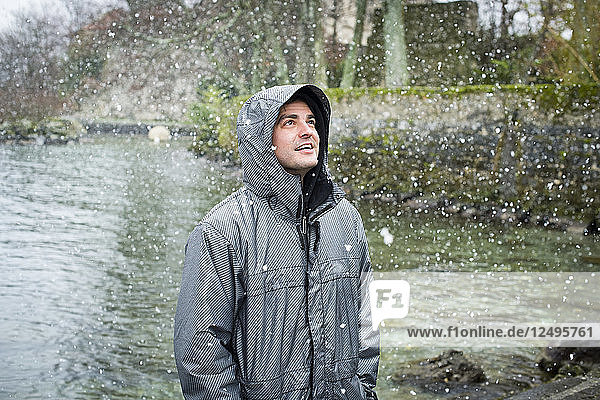 Ein junger Mann in einer Jacke steht an einem See und starrt in den fallenden Schnee in Yvoir  Frankreich.
