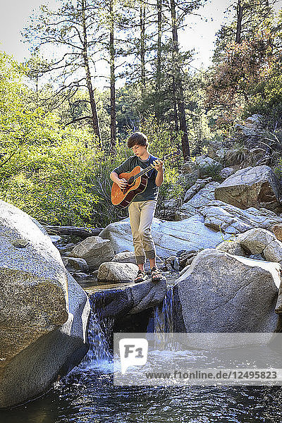 Young man thoughtfully plays his guitar while sitting/standing on the rocks along a creek.