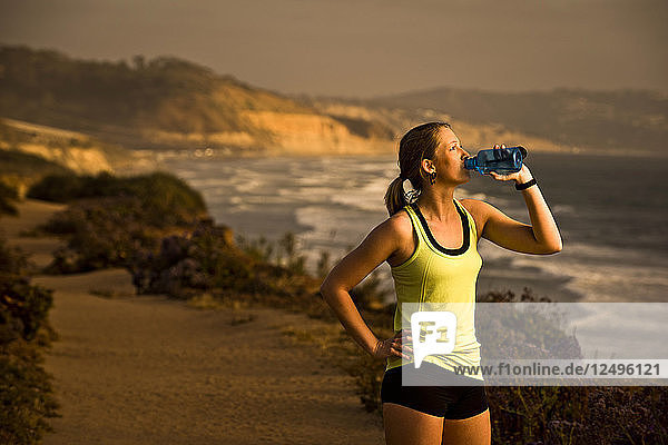 A girl drinks water while taking a break from her run on a trail overlooking the beach at sunset in San Diego California.