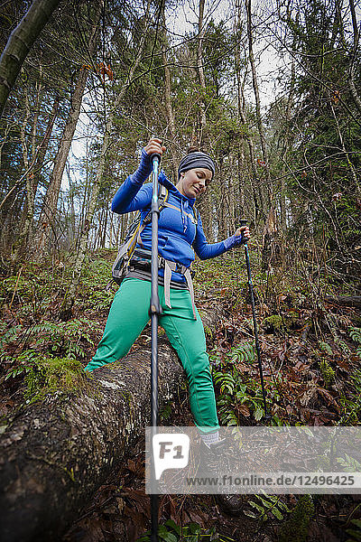 Eine junge Frau benutzt ihre Stöcke  um bei einer Wanderung durch einen dichten Wald einen großen Schritt über einen umgestürzten Baum zu machen.