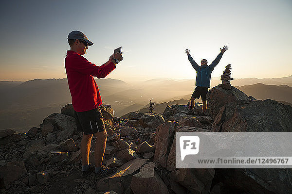 A hiker uses a tablet to take a picture of his friend standing next to the summit cairn of Sauk Mountain  Washington.