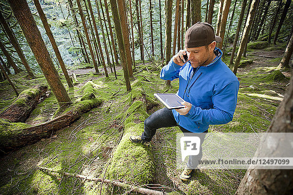 A man talks on phone while working on a tablet in the forest.