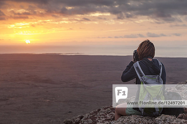 Mädchen mit Rucksack fotografiert den Sonnenuntergang  Sonne fällt ins Meer  von der Spitze eines Berges. Fuerteventura  Kanarische Inseln