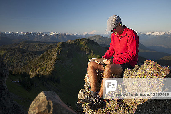 Ein Mann arbeitet an seinem Tablet  während er auf einer felsigen Klippe in der Nähe des Gipfels des Sauk Mountain  Washington  sitzt.