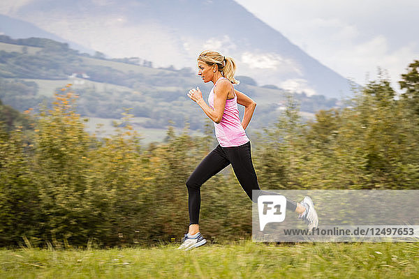 blond woman wearing a pink top and running with a hedge of bushes behind and mountains and clouds in the background in Eloise  France