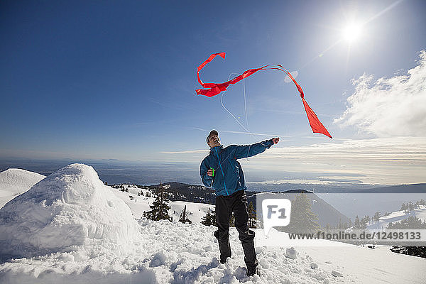 Männlicher Wanderer versucht  einen Drachen auf dem Gipfel eines schneebedeckten Berges steigen zu lassen