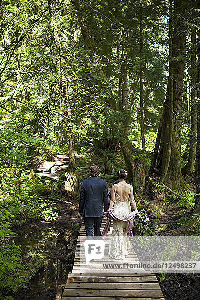 Ein junges Ehepaar hält sich an den Händen  während es über eine Uferpromenade im Wald geht.