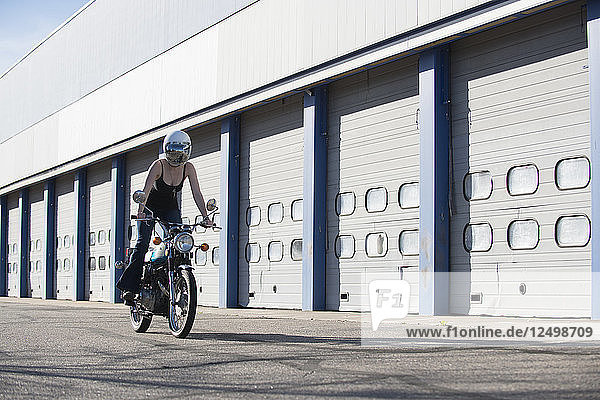 Eine Frau fährt auf ihrem Oldtimer-Motorrad und trägt einen reflektierenden Helm mit Gesichtsschutz  während im Hintergrund eine Reihe von Autogaragentoren zu sehen ist.