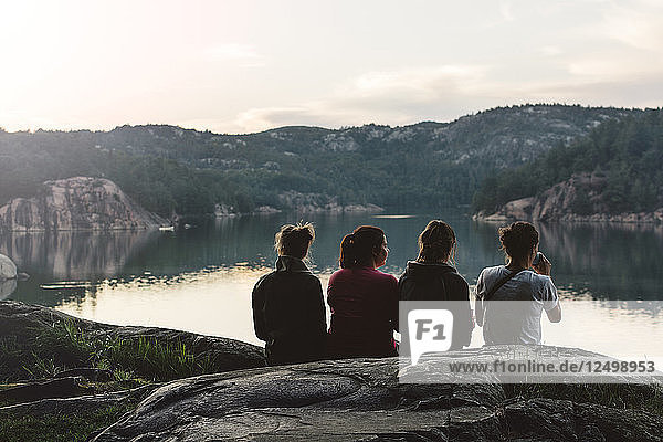 Vier junge Frauen sitzen in der Abenddämmerung am See und entspannen sich