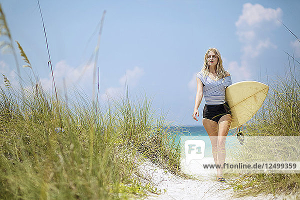 Eine junge Frau geht mit einem Surfbrett einen Weg zum Strand entlang.
