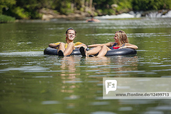 Ein junger Mann und eine Frau lächeln  während sie mit dem Schlauchboot einen Fluss hinunterfahren.