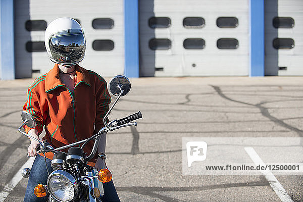 Woman sitting on her vintage motorcycle wearing a reflective helmut face shield with a line of auto garage doors in the background.
