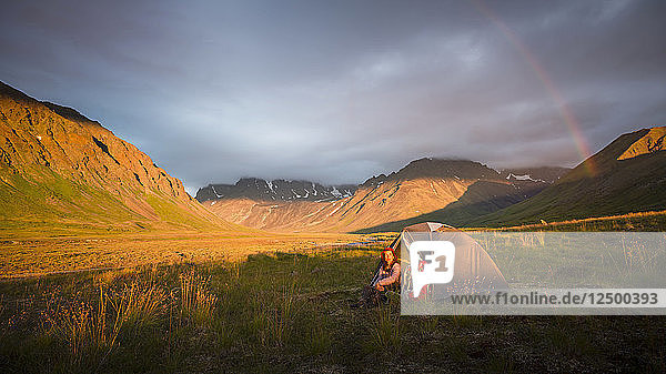 Eine Frau sitzt bei ihrem Zelt  als ein Regenbogen über dem Camp im Lake Clark National Park and Preserve aufzieht