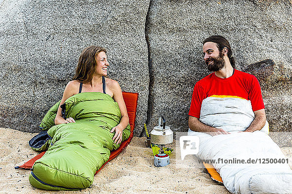 Mann und Frau am Strand in Schlafsäcken  lachend mit einem Kocher  der Wasser erhitzt.