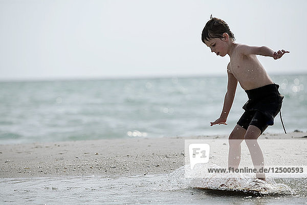 A young boy in a wetsuit surfs on his skim board at the shore.