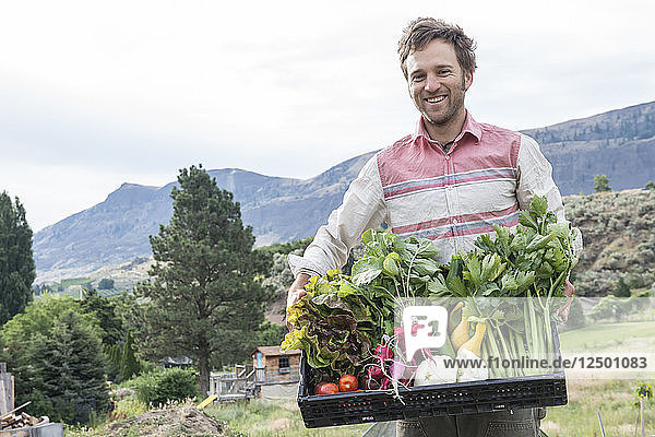 Ein Mann hält eine Kiste mit frisch geerntetem Obst und Gemüse auf einem Biohof