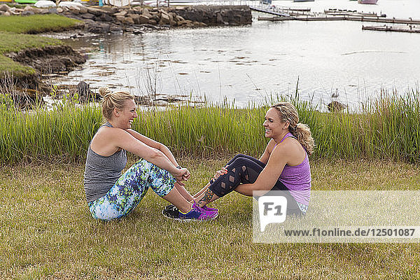 Two female friends sitting on the grass laughing near the water.