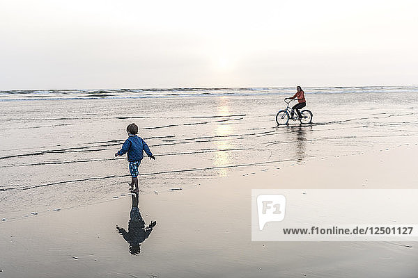 Eine Frau fährt mit dem Fahrrad  während ihr kleiner Junge am Strand spielt.