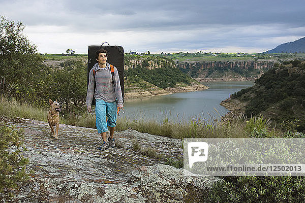 A Male Climber Carrying A Crash Pad Explores The Surroundings Of A Lake With His Dog