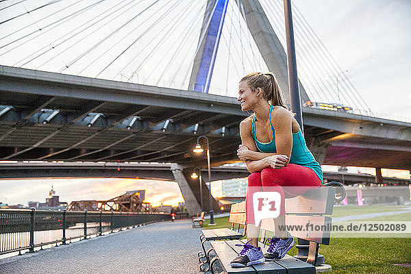 Frau lächelnd und auf einer Bank sitzend in Trainingskleidung bei Sonnenuntergang in einem Park in der Nähe einer Brücke.