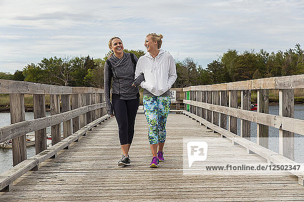 Zwei Freundinnen auf einem Steg in der Nähe des Wassers lachen  während sie trainieren.