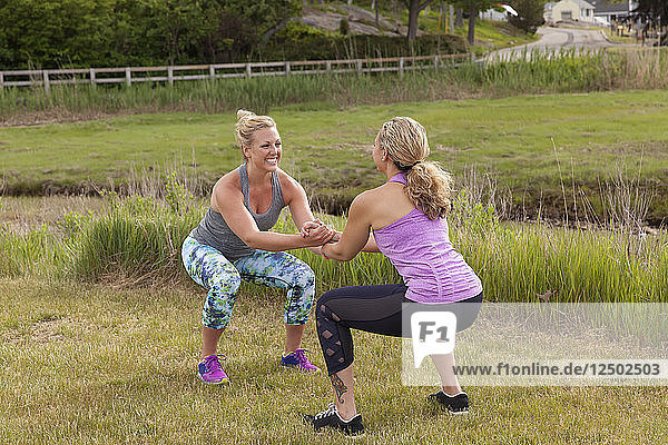 Zwei Frauen machen Kniebeugen in einem Partner-Workout draußen auf dem Rasen am Wasser.