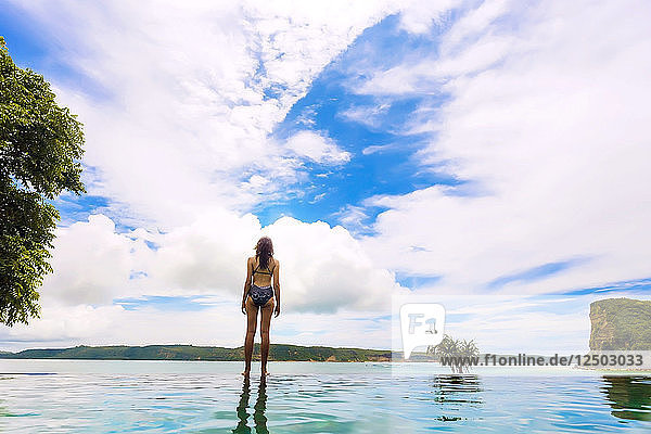 Rear view of woman in bikini standing on lakeshore against white clouds