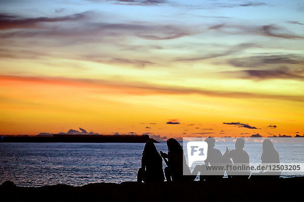 Silhouetten von Menschen sitzen am Meeresufer bei Sonnenuntergang