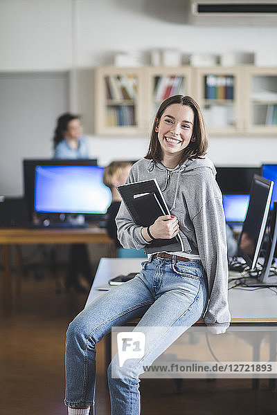 Cheerful high school female student sitting with books on desk in classroom